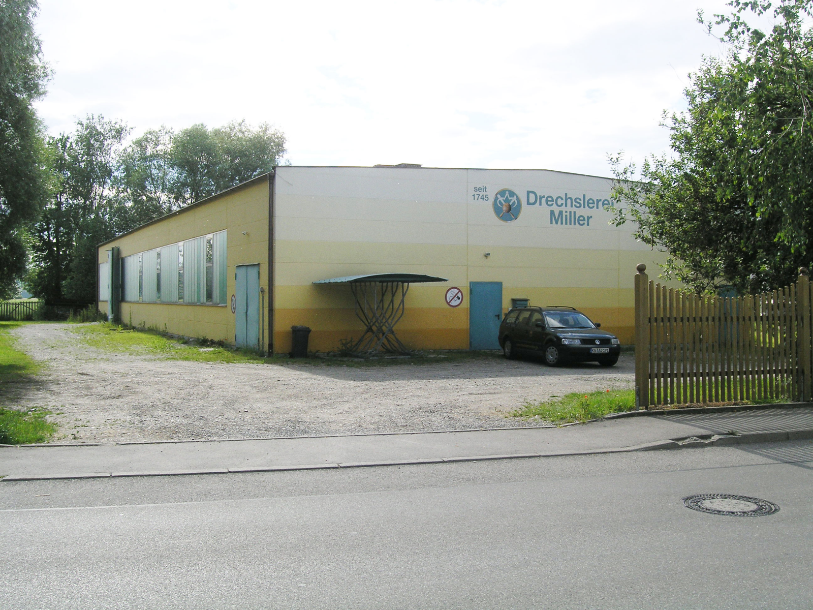 Aufnahme des 1972 errichteten Betriebsgebäudes der Drechslerei Miller im Industriegebiet: gelb-weiß gestrichene Halle mit seitlichen Fensterbändern und Firmenlogo.